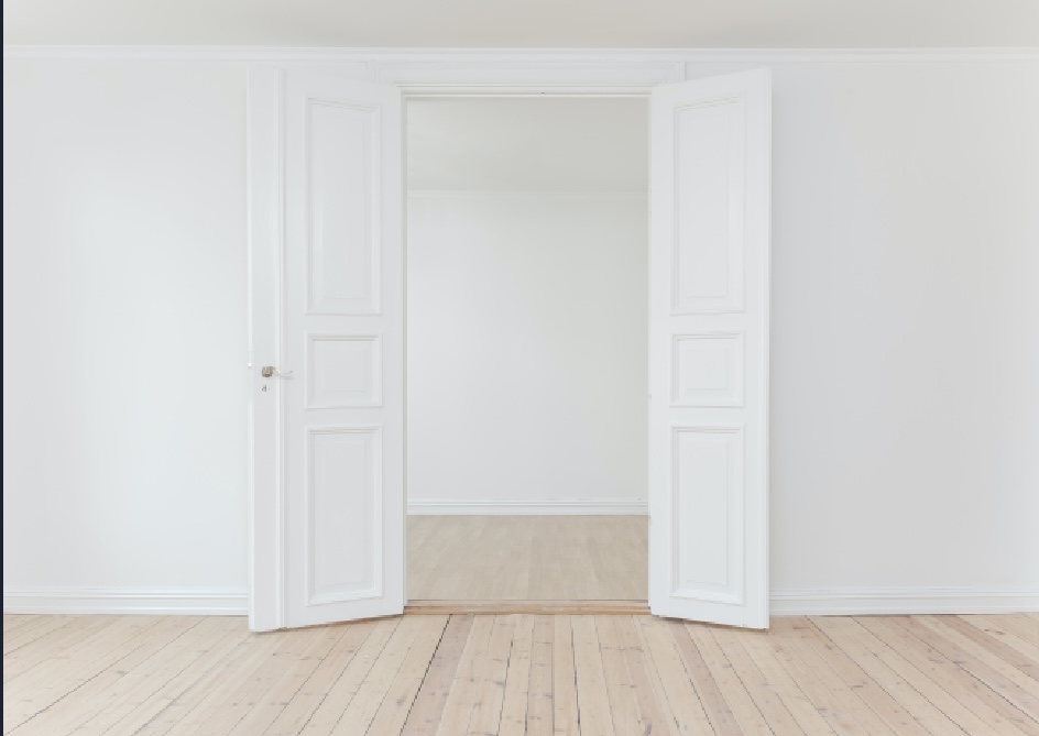 A bright photo of two white doors open between two rooms with white walls and birch flooring.