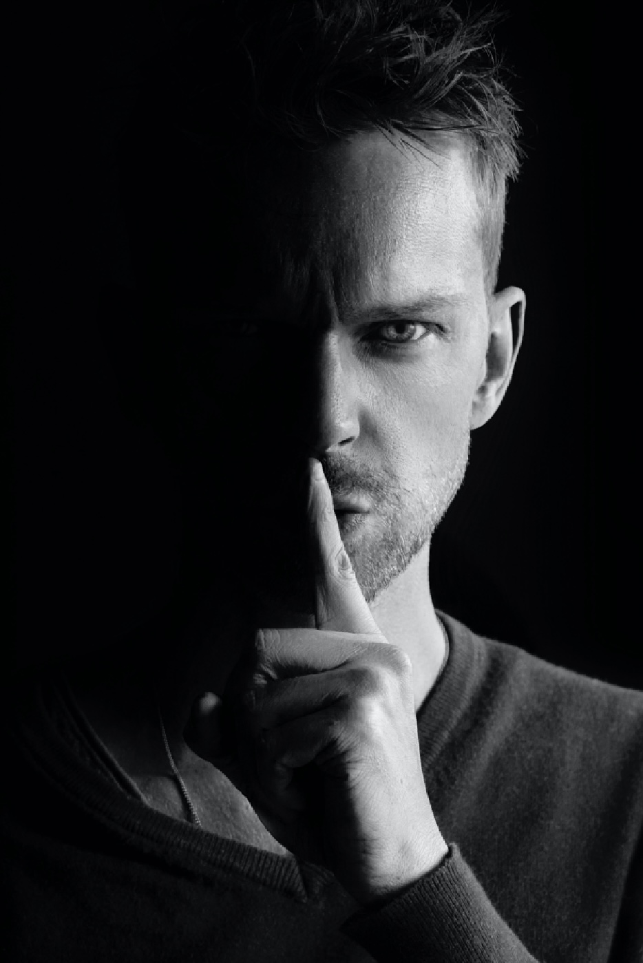 A black and white photo of a man covering his lips with his finger as if saying “ssshhh”.