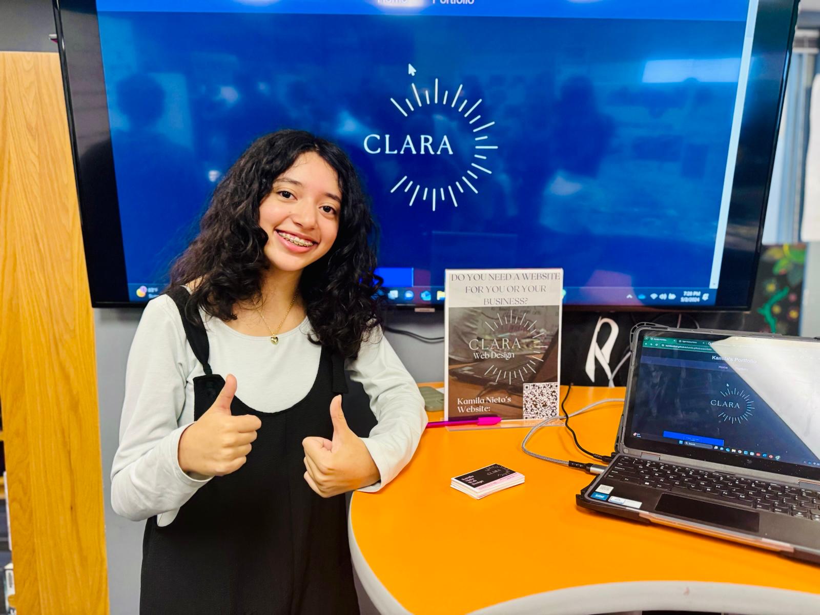 Me at my station at the entreprenuership fair in front of a TV and laptop that both say 'Clara'.