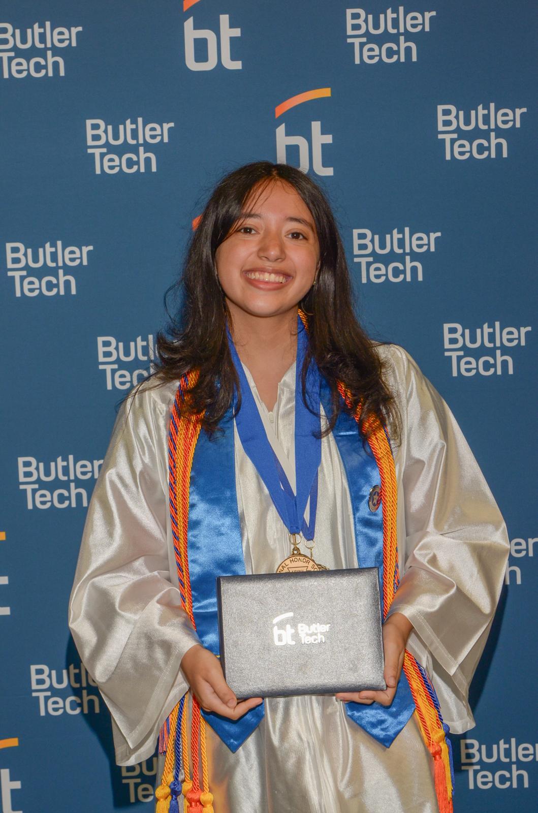 Me in my graduation gown in front of a Butler Tech background, holding my certificate of completion.